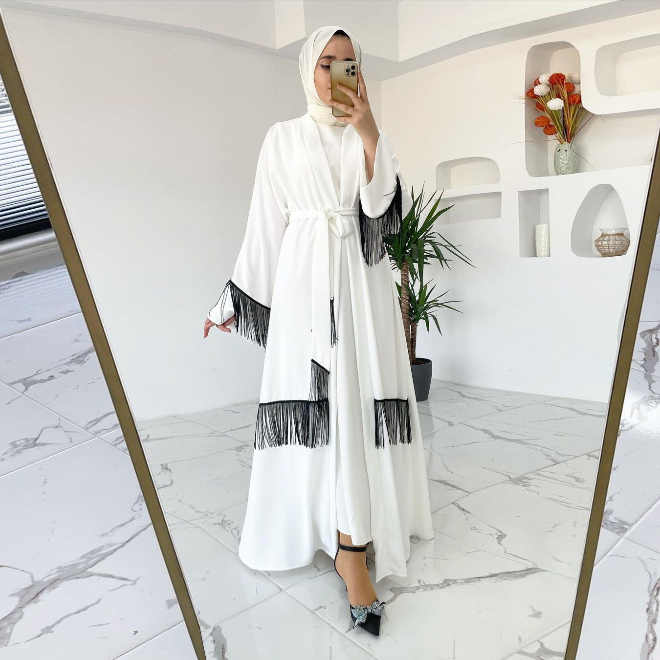 Woman taking a mirror selfie wearing a white abaya with black fringe details in a modern interior setting.