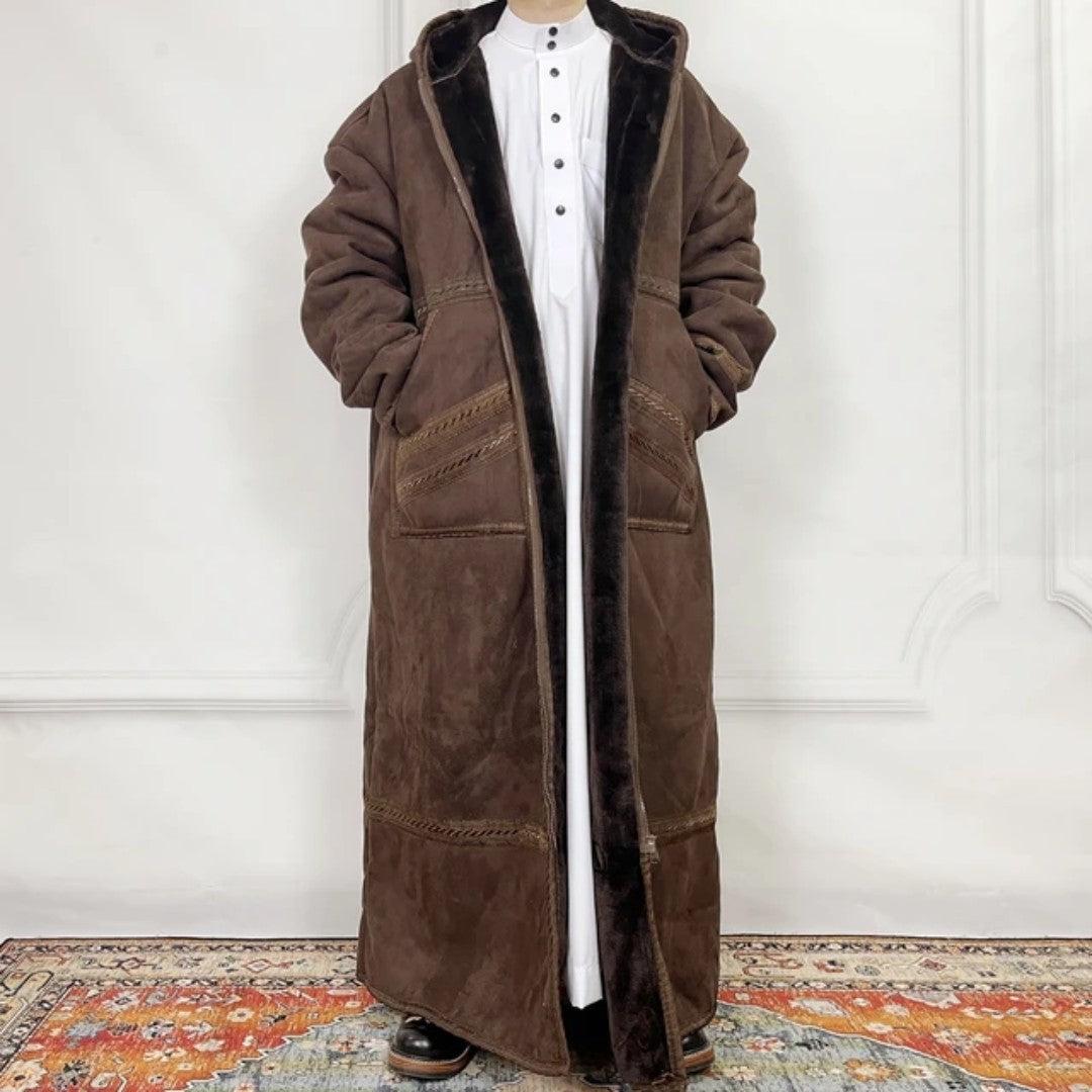 Brown long coat with fur lining worn by a person against a white wall.