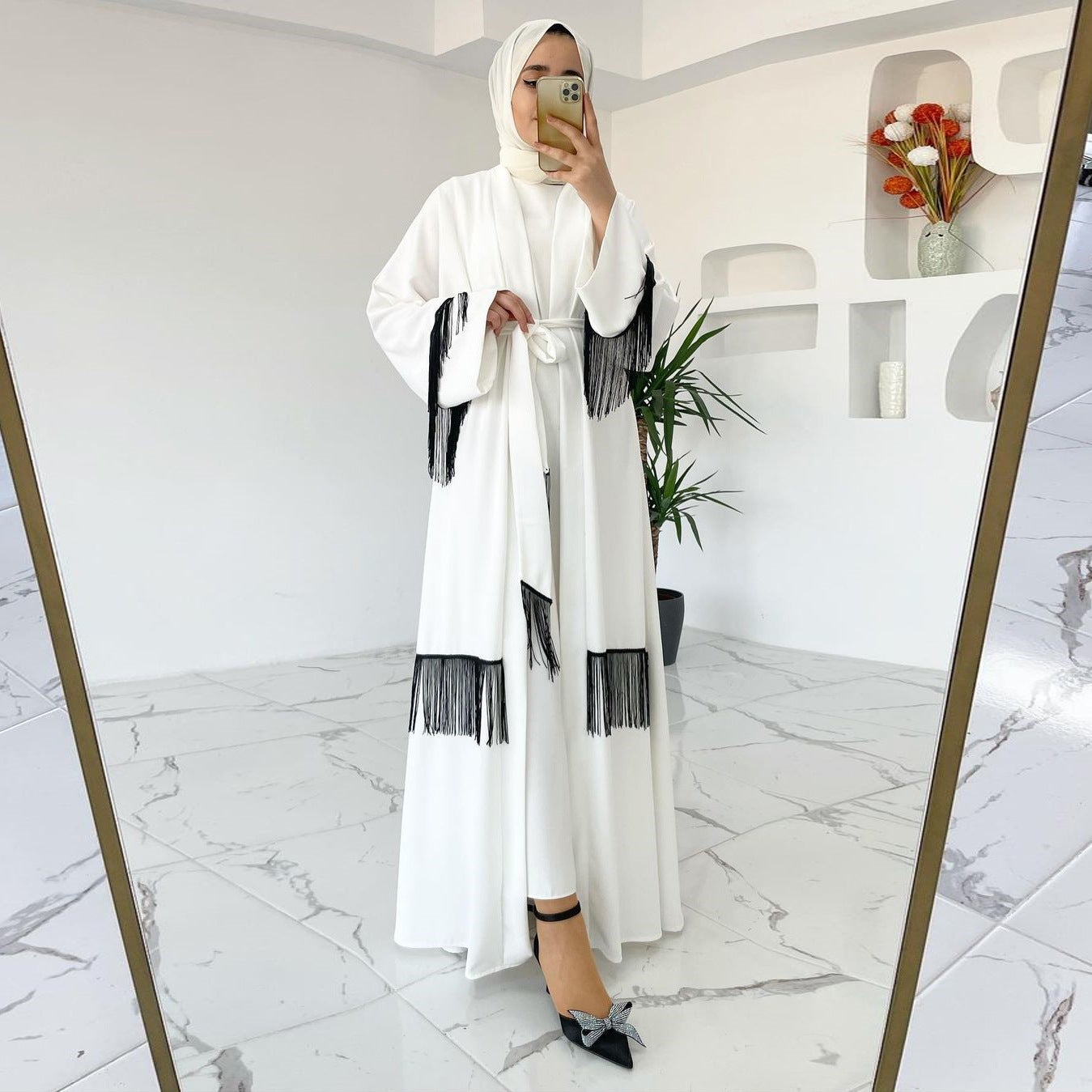 Woman in a white abaya with black details taking a mirror selfie in a modern interior setting.