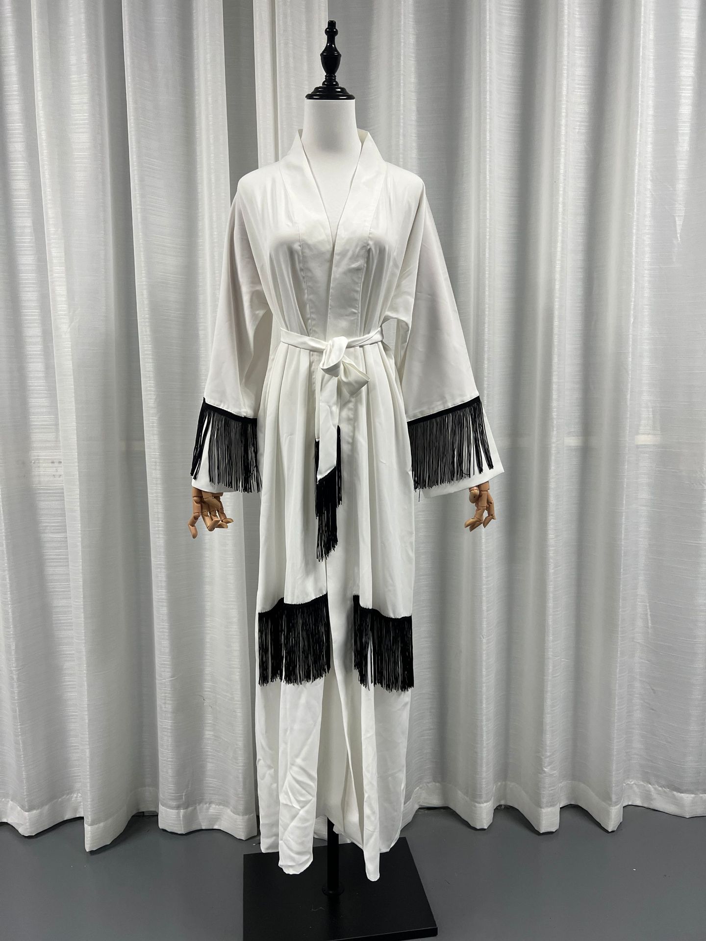 White robe with black fringe details on a mannequin against a white curtain background