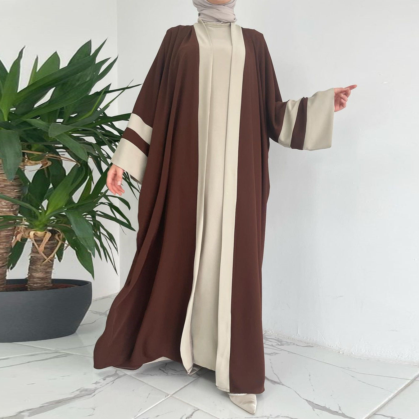 Person wearing a brown and beige abaya standing next to a potted plant on a white floor.