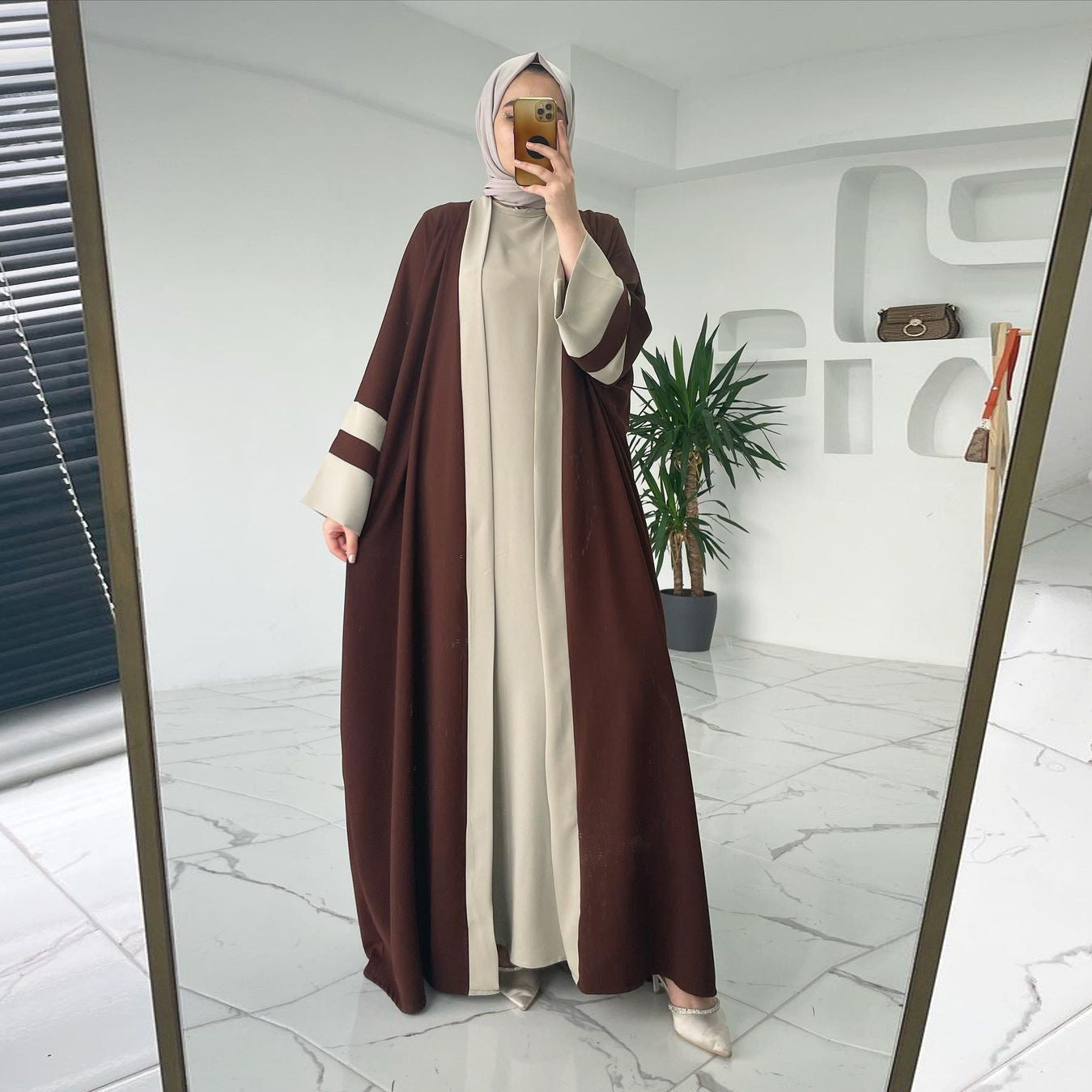 Person wearing a brown and beige abaya taking a mirror selfie in a modern interior setting.