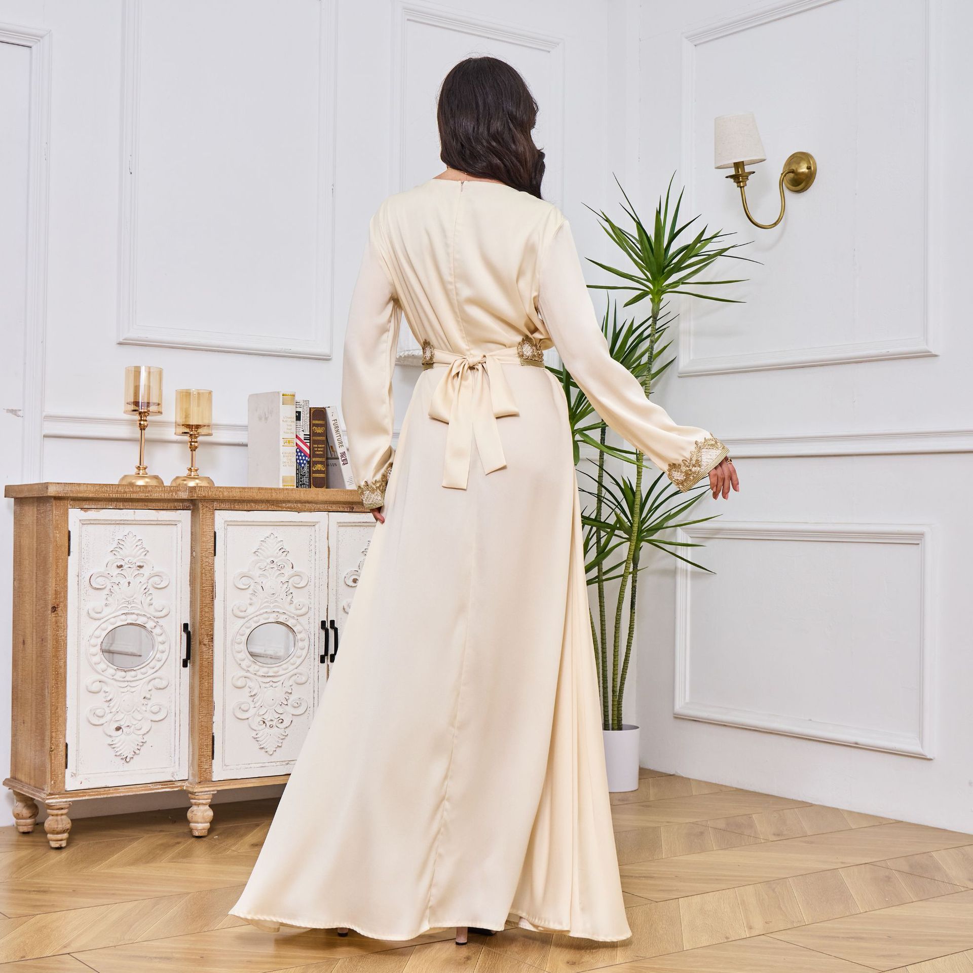 Woman in a long beige dress standing in a room with white walls and wooden flooring.